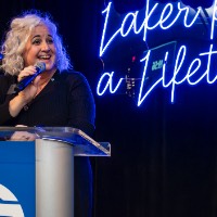 Susan Proctor speaks into microphone on podium in front of Laker for a Lifetime neon sign.